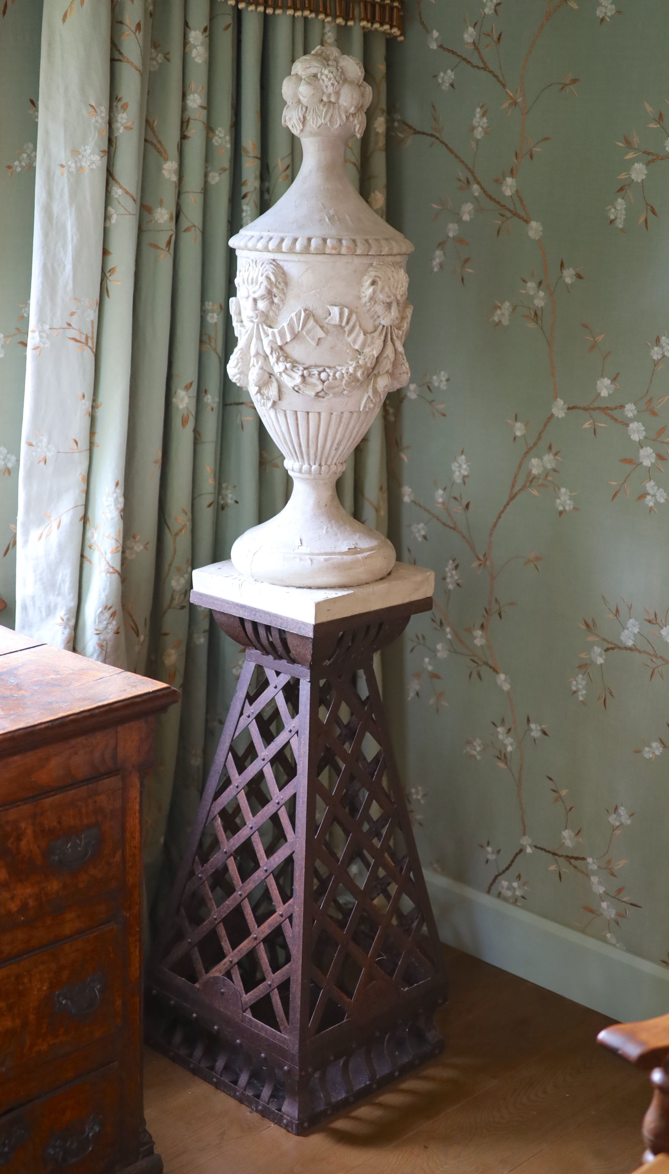 A pair of white painted carved walnut urns and covers, on wrought iron stands, the urns carved with fruit, lions masks and floral swags, height 90cm, the tapered stands with lattice work sides, height 90cm, overall heigh